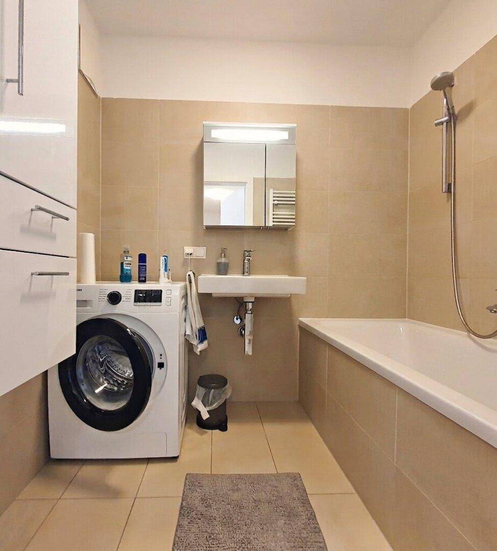 Modernes Badezimmer mit Badewanne, Waschmaschine und beigefarbenen Fliesen.