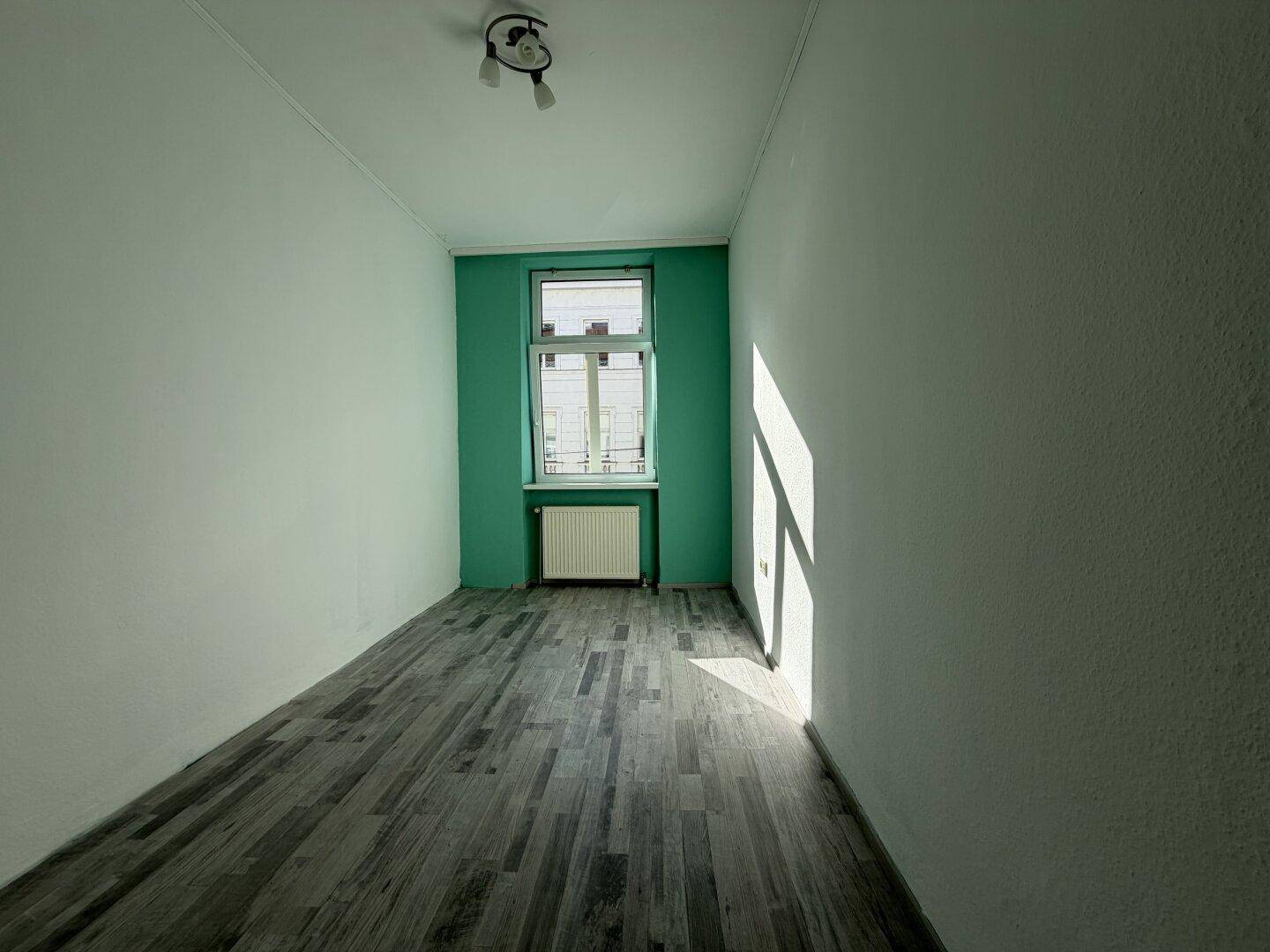 Heller Raum mit dunklem Laminatboden und einer farbigen Akzentwand um das Fenster, durch das Sonnenlicht fällt.