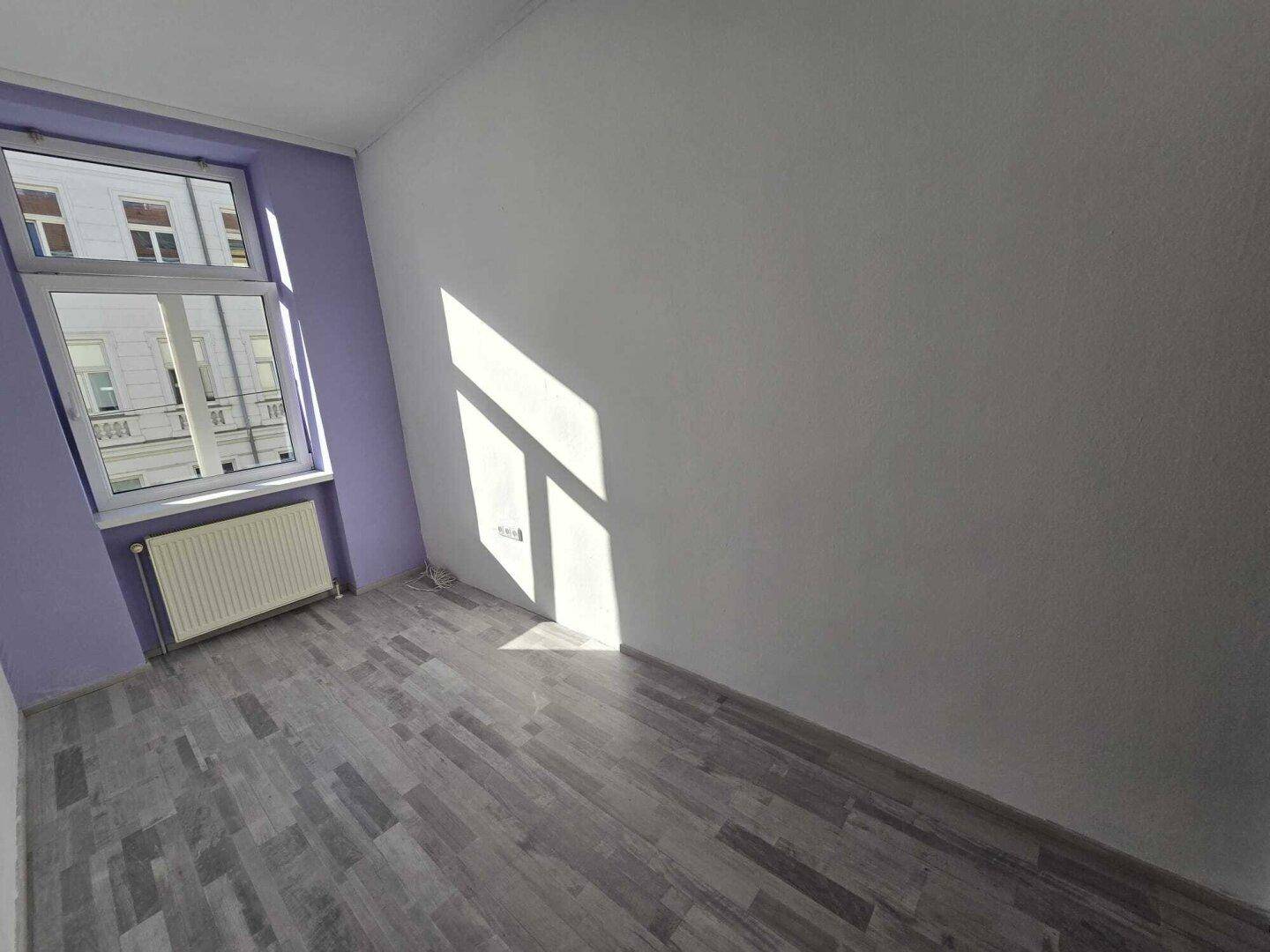 Heller Raum mit grauem Laminatboden und einer lila Akzentwand um das Fenster, durch das Sonnenlicht fällt.