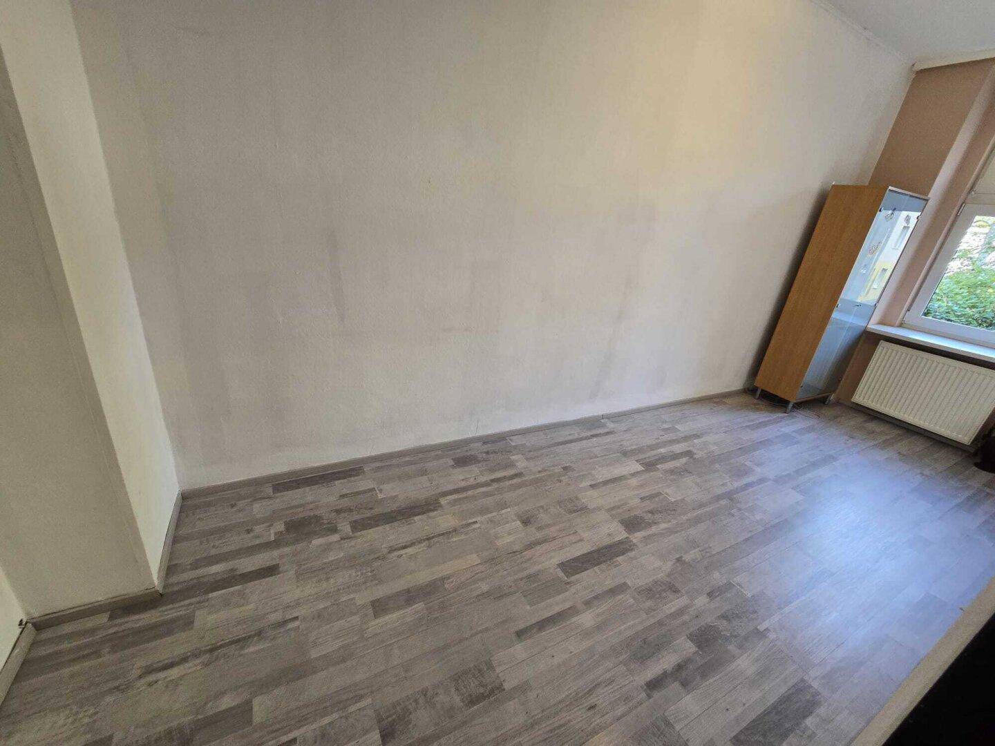 Heller, leerer Raum mit grauem Laminatboden und einem Fenster, das Tageslicht hereinlässt.