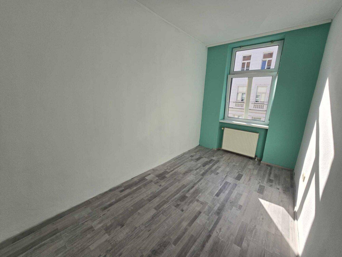 Leeres Zimmer mit dunklem Laminatboden und einer auffälligen türkisfarbenen Wand um das Fenster.
