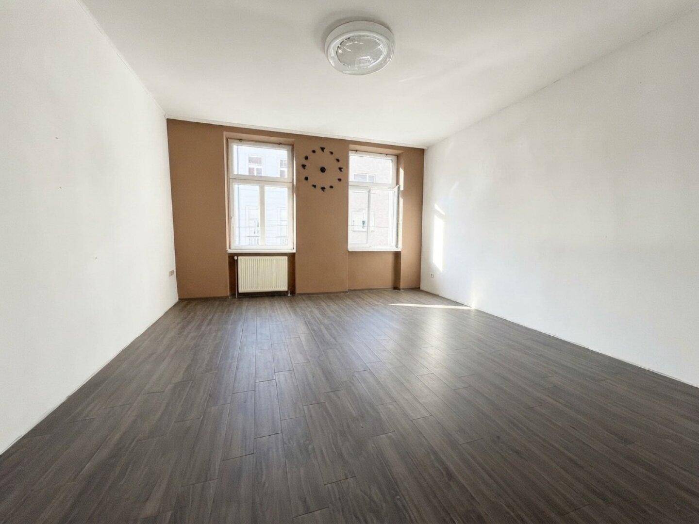 Helles, leeres Zimmer mit dunklem Laminatboden, einer braunen Akzentwand und zwei Fenstern.