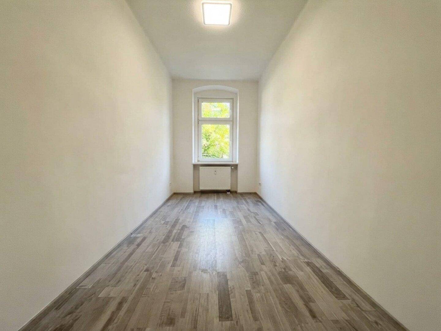 Schmaler, heller Raum mit dunklem Laminatboden und einem Fenster mit Blick ins Grüne.