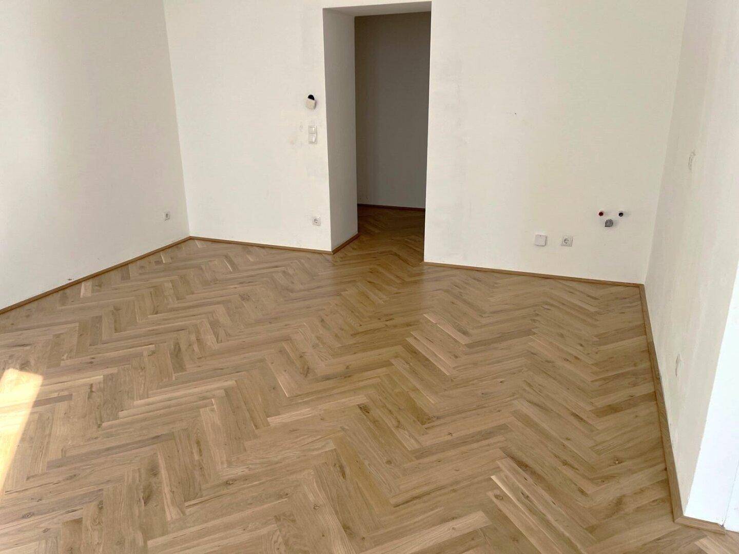 Leerer Wohnraum mit elegantem Fischgrätparkett und weißen Wänden, bereit zum Einrichten.