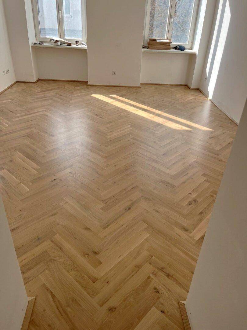 Großer, lichtdurchfluteter Raum mit glänzendem Fischgrätparkett und zwei Fenstern.