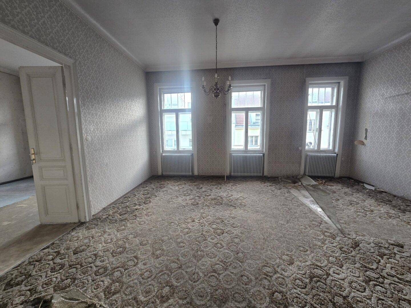 Geräumiges Wohnzimmer mit altem Teppichboden, Tapeten und drei großen Fenstern, renovierungsbedürftig.