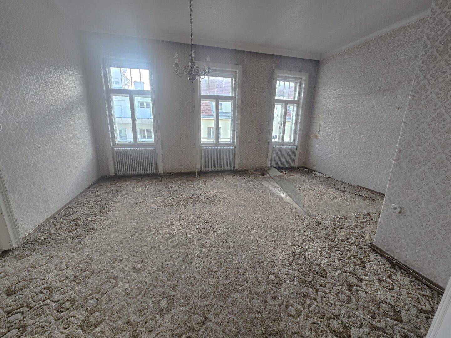 Helles Wohnzimmer mit altem Teppichboden und Tapeten, bereit für eine Modernisierung.