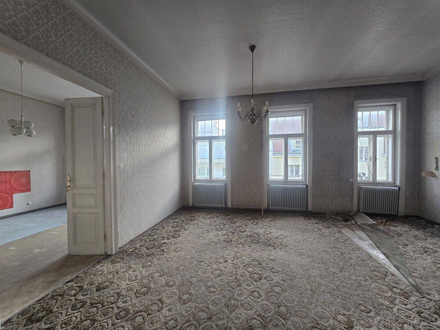 Großes Wohnzimmer mit Teppichboden und Zugang zu einem weiteren Raum, viel Tageslicht.
