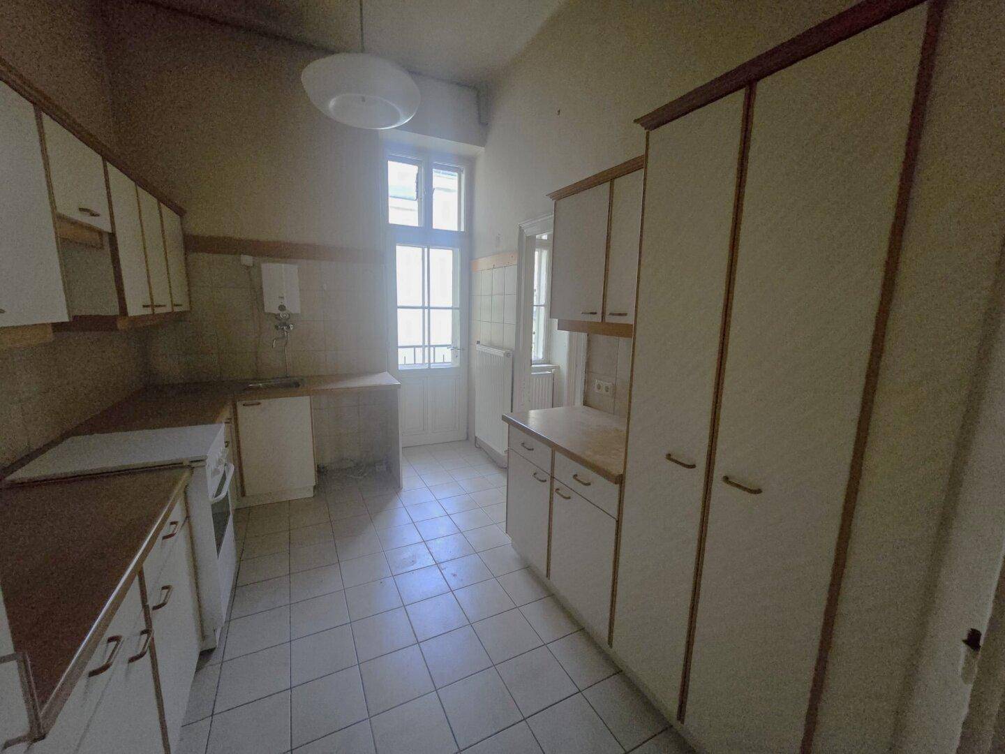 Küche im Originalzustand mit weißen Schränken, Fliesenboden und Fenster, bereit zur Modernisierung.