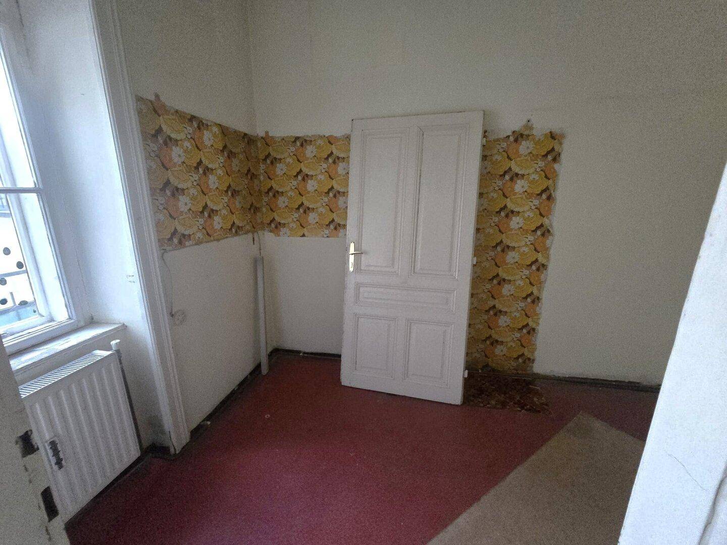 Kleines Zimmer mit floraler Tapete, rotem Teppichboden und Fenster, renovierungsbedürftig.