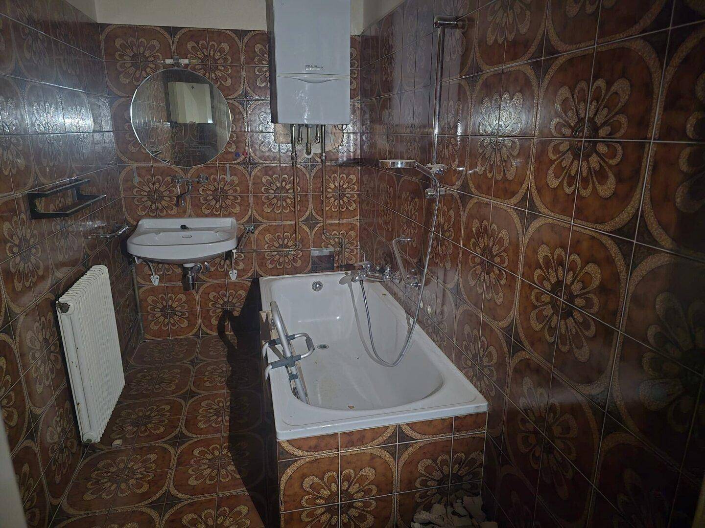 Badezimmer im Originalzustand mit braunen gemusterten Fliesen, Badewanne und Waschbecken.