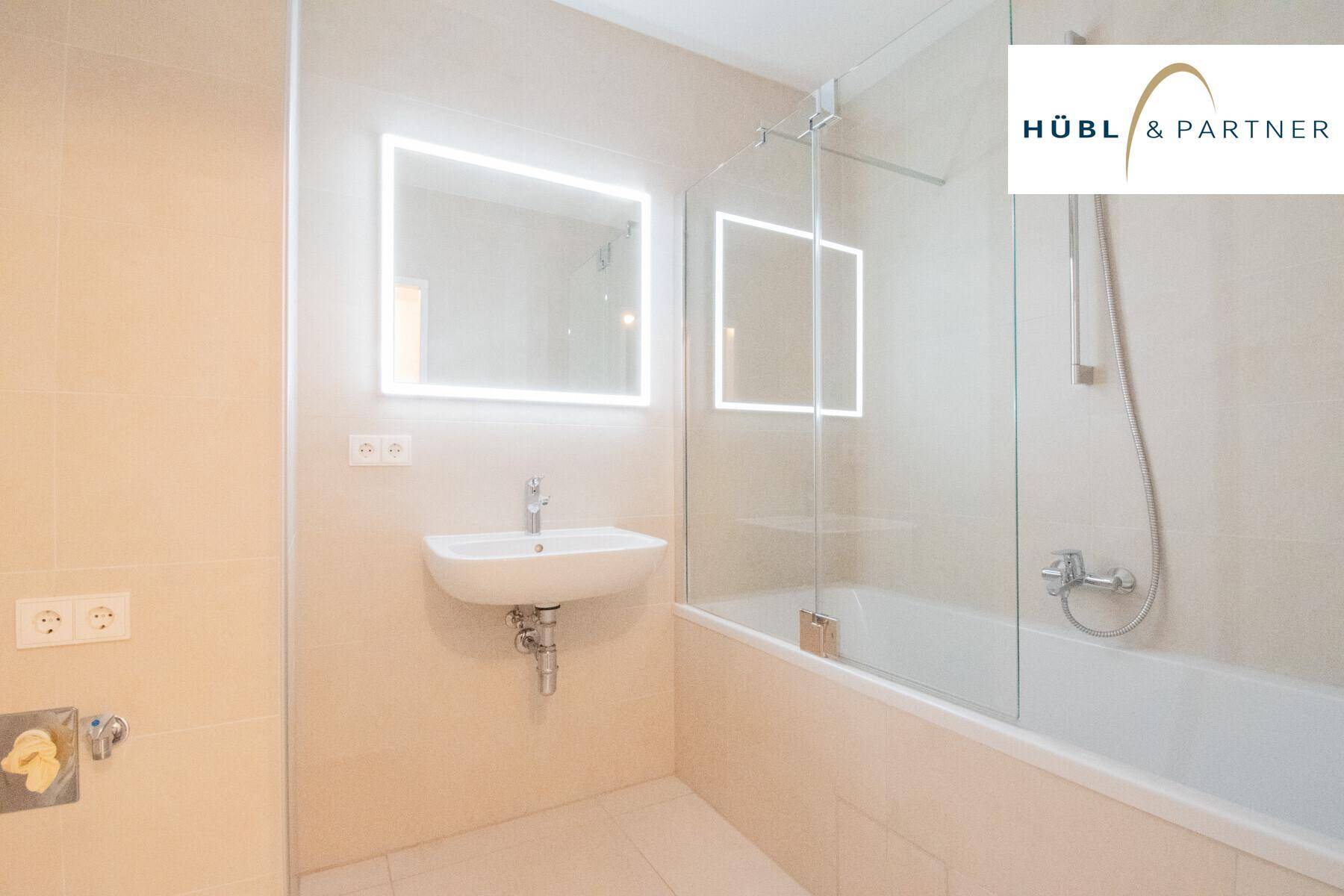 Modernes Badezimmer mit beigen Fliesen, beleuchtetem Spiegel und einer Badewanne mit Duschwand.
