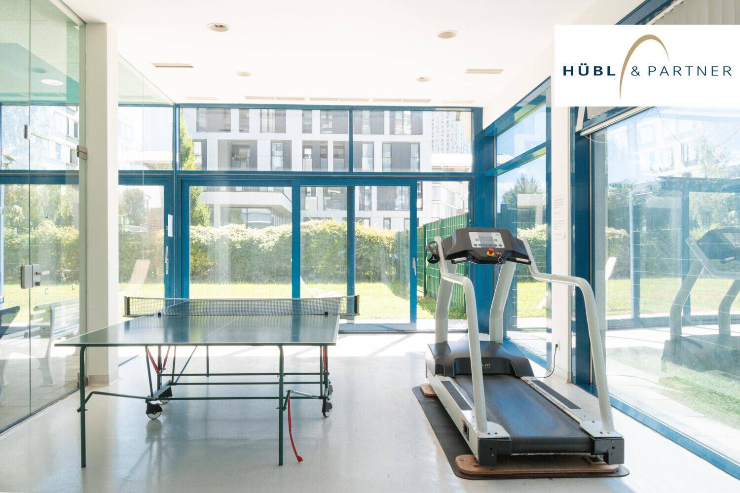 Fitnessbereich mit Laufband und Tischtennisplatte, umgeben von großen Glasfenstern.