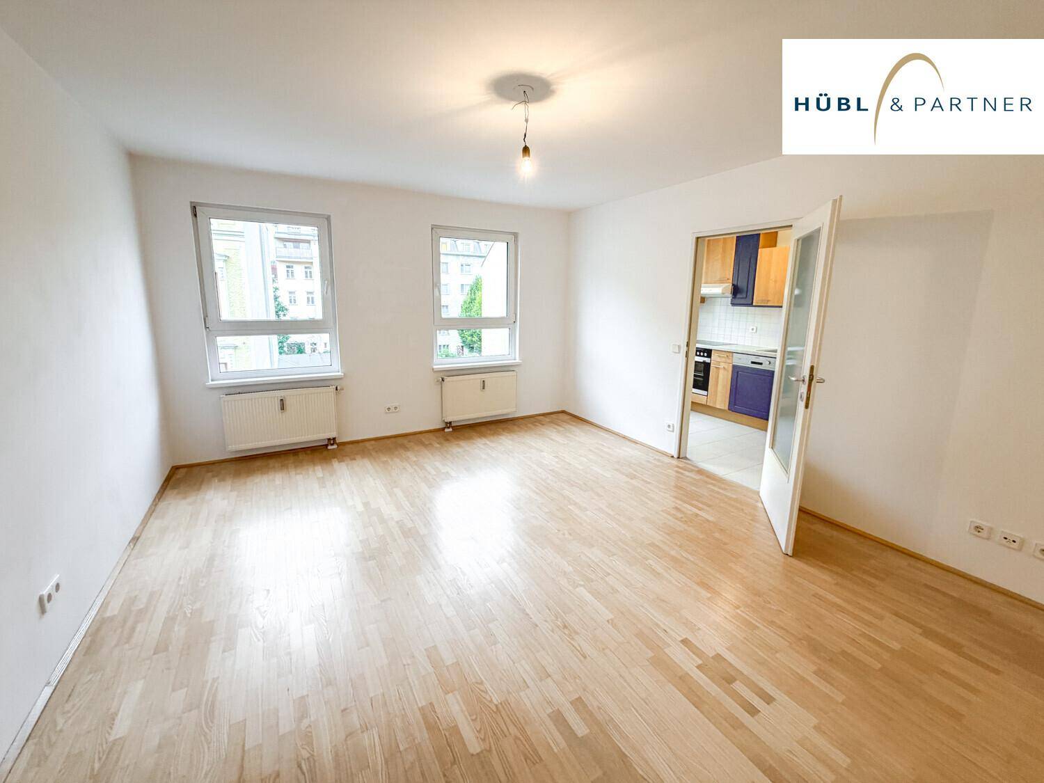 Helles, leeres Wohnzimmer mit Parkettboden und Zugang zu einer separaten Küche.