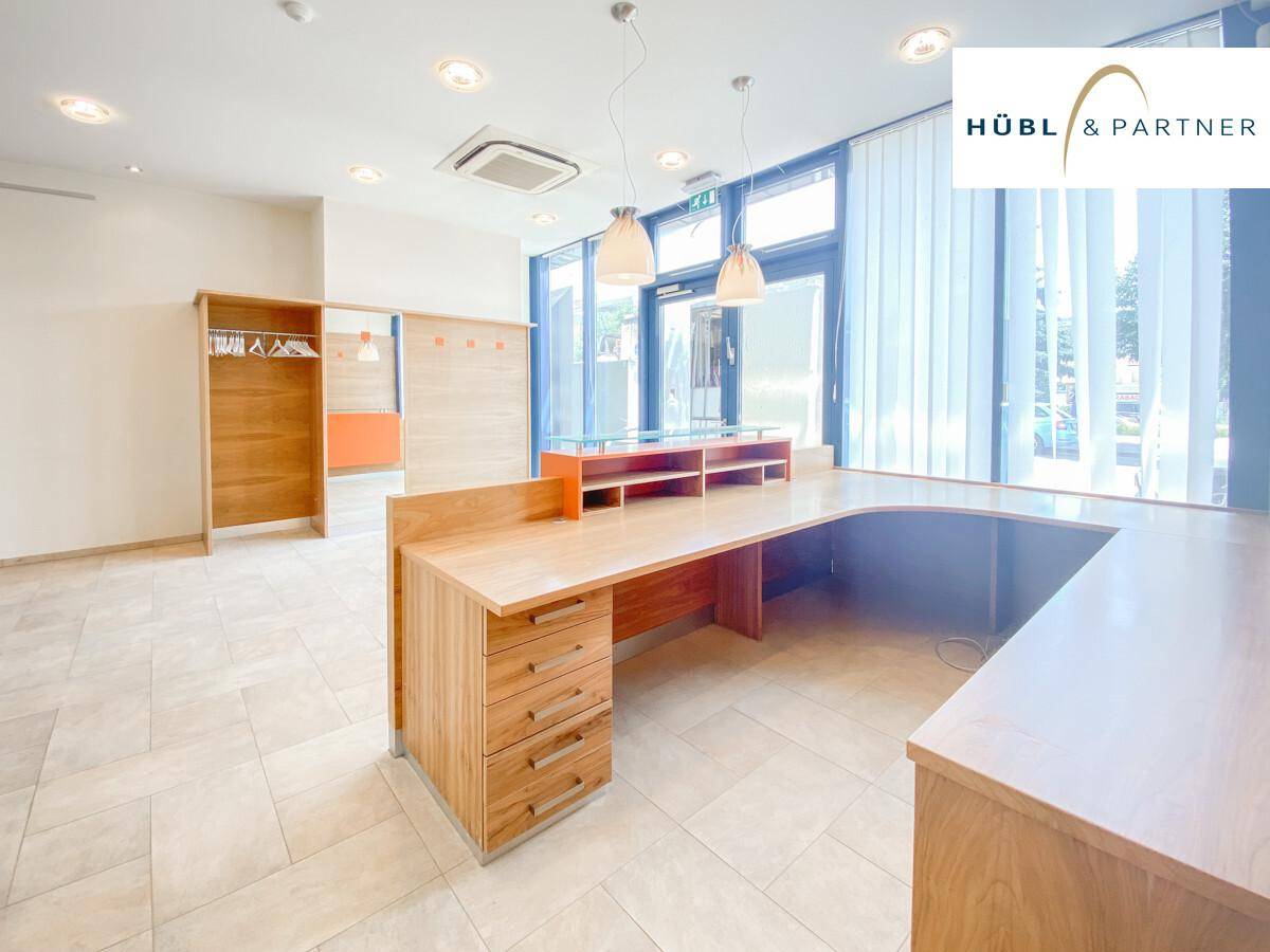Heller Bürobereich mit großem Holzschreibtisch, Garderobe und Blick durch die Fenster auf den Außenbereich.
