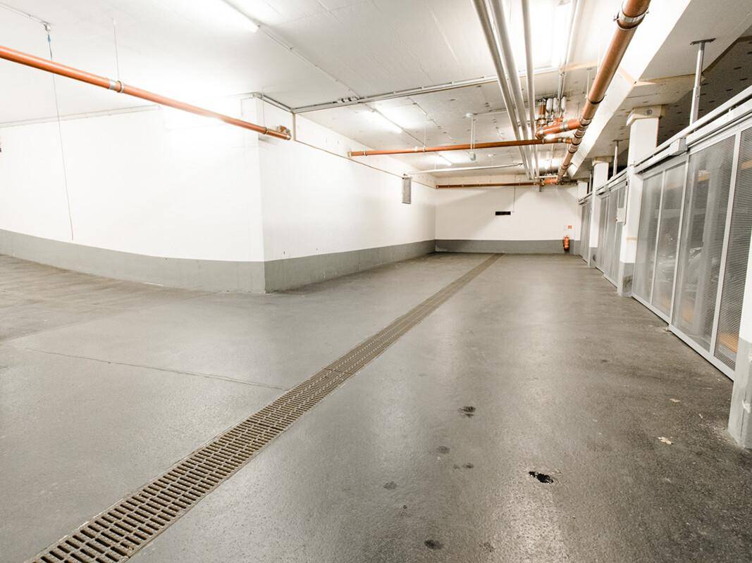 Geräumige Tiefgarage mit Betonboden, weißen Wänden und sichtbaren Rohrleitungen an der Decke.