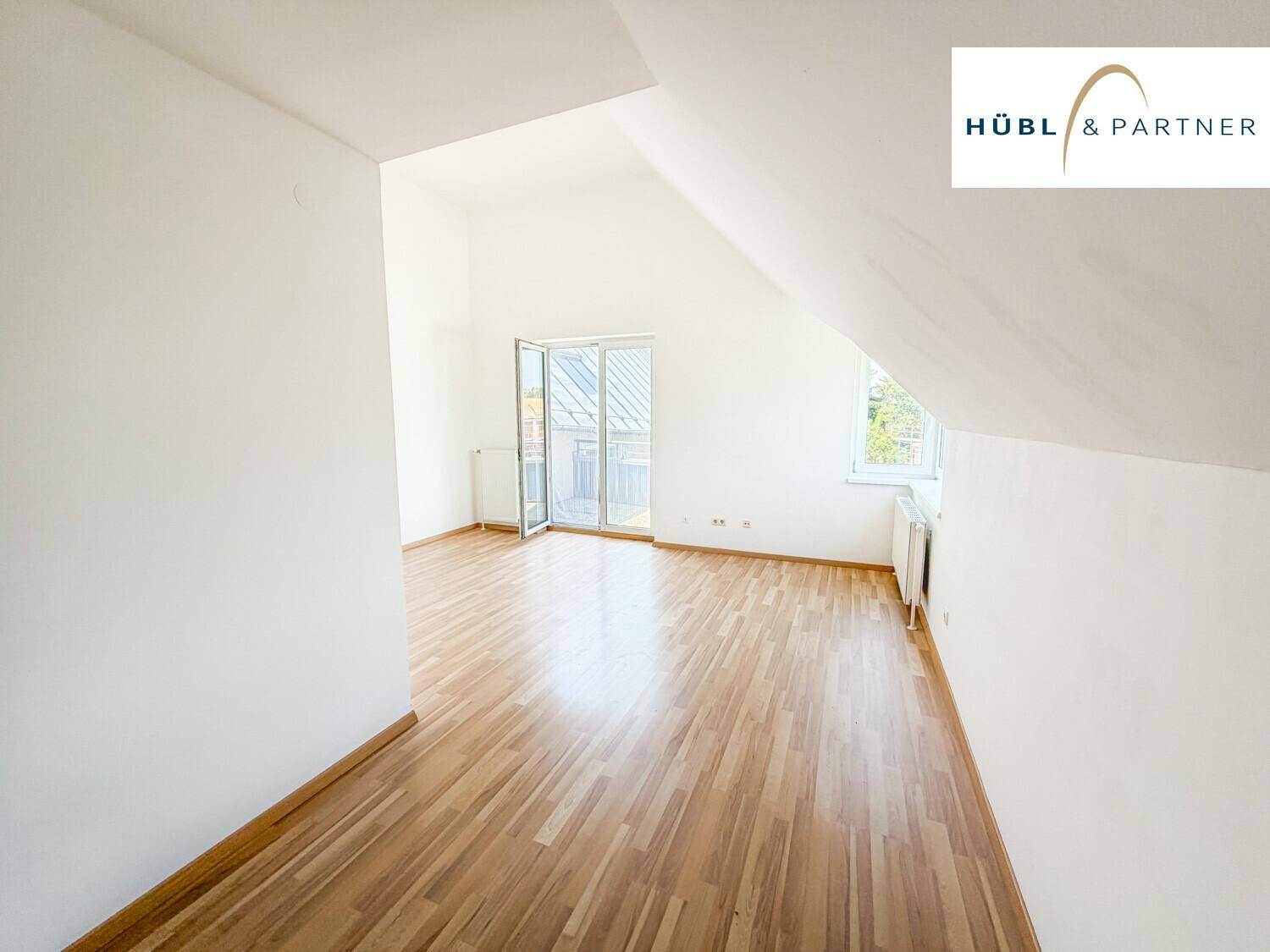 Heller Wohnraum im Dachgeschoss mit Holzboden, weißen Wänden und Zugang zu einem Balkon.