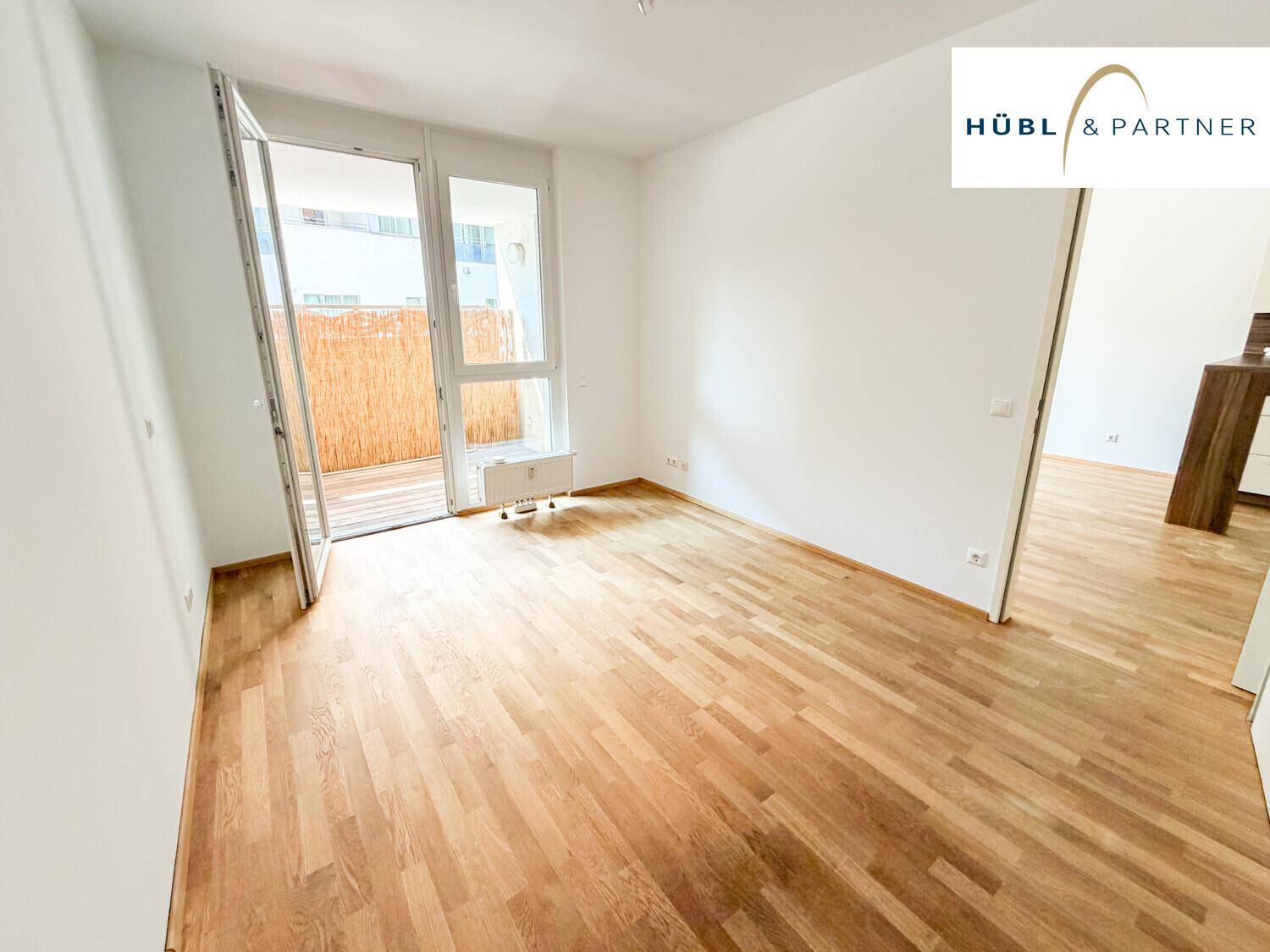 Geräumiger, heller Raum mit Holzboden und großem Balkonzugang, der viel Tageslicht hereinlässt.