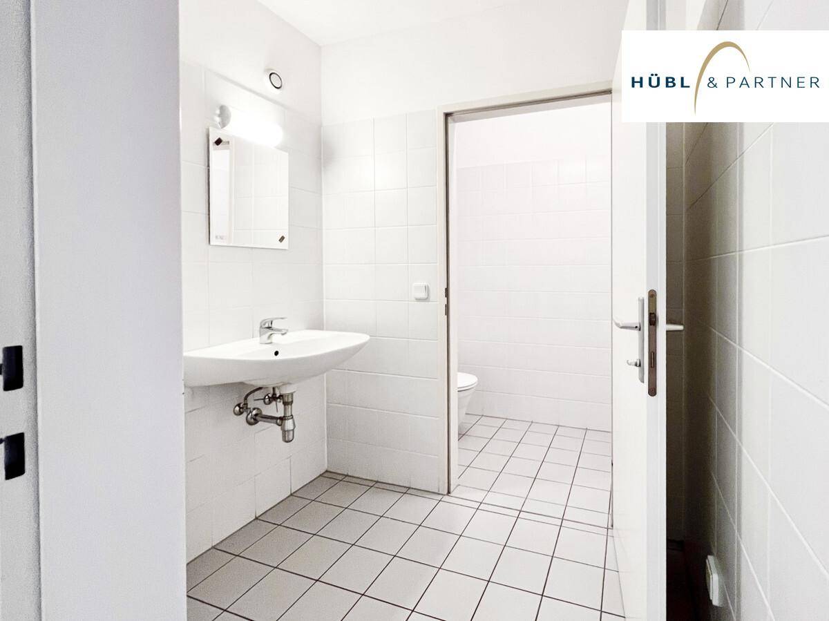 Helles Badezimmer mit weißen Fliesen, einem Waschbecken und einer Toilette, die durch die offene Tür sichtbar ist.
