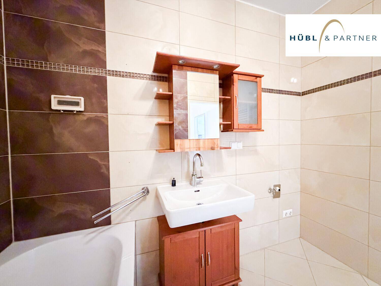 Badezimmer mit Einzelwaschbecken, Holzunterschrank und Spiegelschrank, umgeben von hellen Fliesen.
