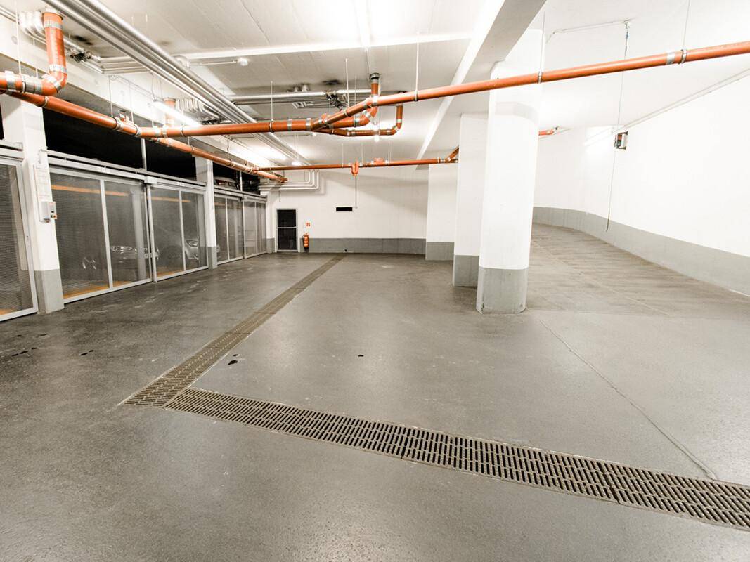 Weitläufige Tiefgarage mit Betonsäulen, Gitterboxen und sichtbaren Installationen an der Decke.