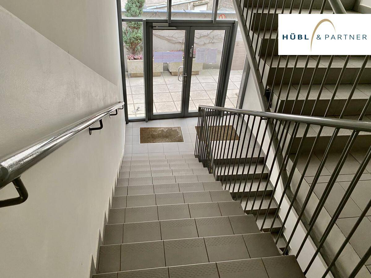 Treppenhaus mit Fliesenboden und Metallgeländer, das zu einem Ausgang mit Glastüren im Erdgeschoss führt.