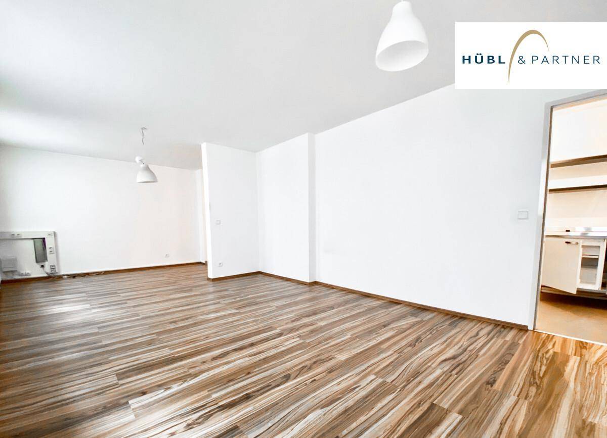 Heller Wohnraum mit Holzfußboden und Zugang zu einem angrenzenden Bereich, möglicherweise einer kleinen Küche.