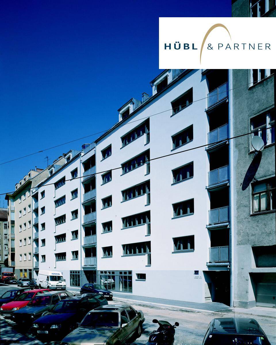 Mehrstöckiges Wohngebäude mit weißer Fassade und vielen Fenstern, gelegen an einer Straße mit parkenden Autos.