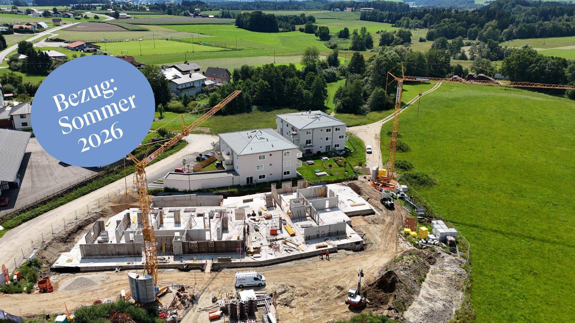 Luftaufnahme einer Baustelle mit neuen Gebäuden und grüner Umgebung, Bezug Sommer 2026.