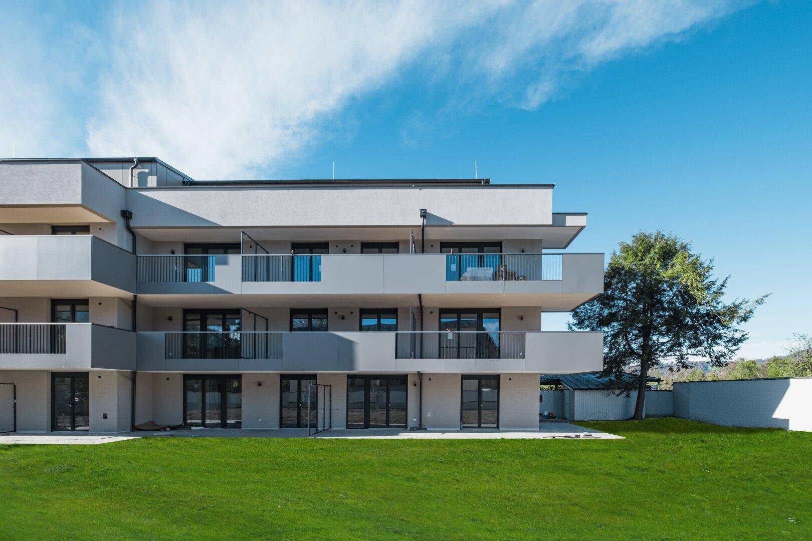 Seitenansicht des modernen Wohngebäudes mit Balkonen und weitläufiger Rasenfläche unter blauem Himmel.