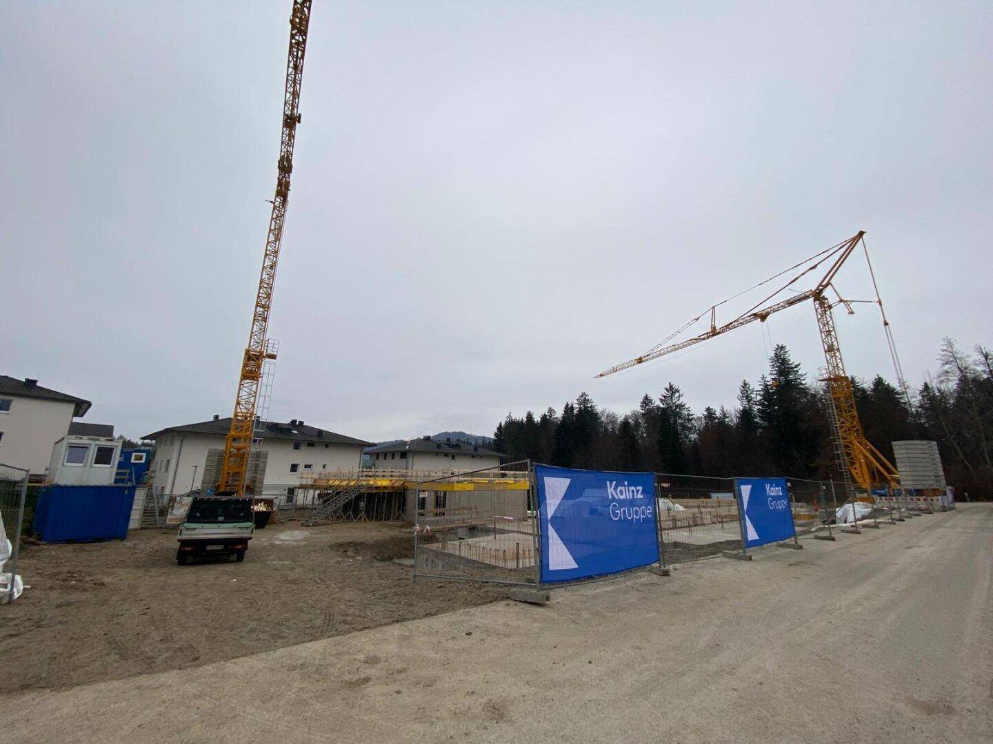 Weitläufige Baustelle mit zwei Kränen und Werbebannern, umgeben von Bäumen und Wohngebäuden.