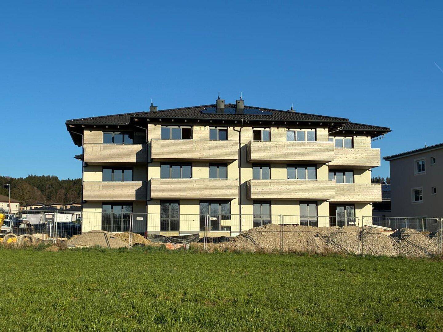 Frontansicht eines im Bau befindlichen Mehrfamilienhauses mit Balkonen und Solarpaneelen auf dem Dach.