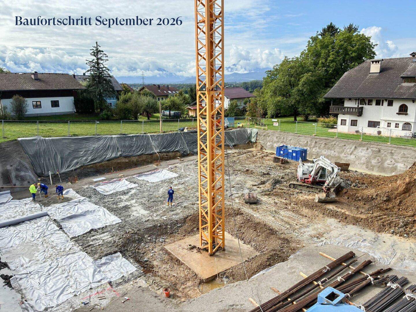 Baufortschritt einer Baustelle mit ausgehobener Grube, Kran und Bauarbeitern. Im Hintergrund sind Wohnhäuser und Berge sichtbar.