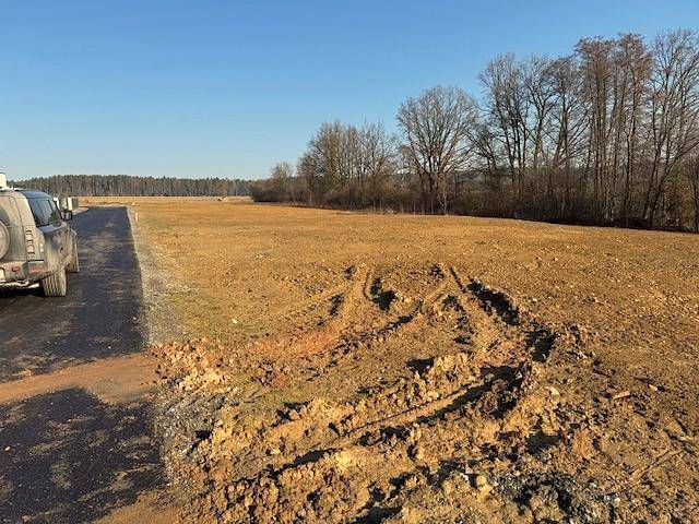 Großes, unbebautes Grundstück neben einer asphaltierten Straße mit einem Fahrzeug und Wald im Hintergrund.