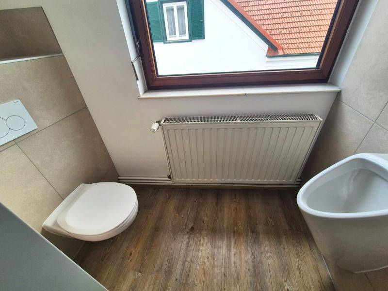 Badezimmer mit Toilette, Urinal und einem Fenster, das einen Blick auf die umliegenden Dächer bietet.