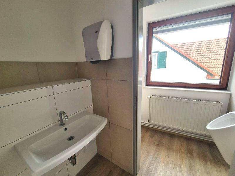 Badezimmer mit Waschbecken, Fliesenwand und einem Fenster mit Blick auf ein Ziegeldach und Nachbargebäude.