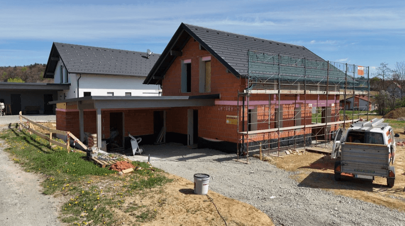 Einfamilienhaus im Rohbau mit Carport und Gerüst auf einem sonnigen Grundstück.