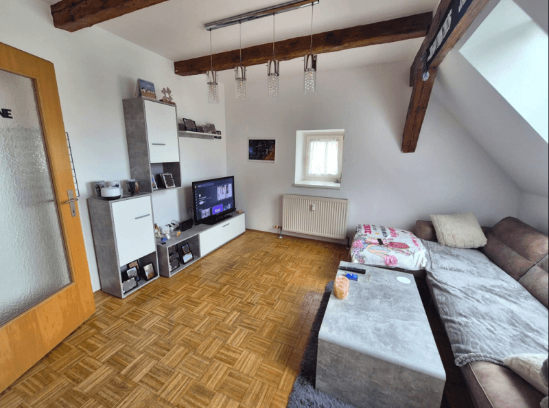 Helles Wohnzimmer mit Holzbalken, Parkettboden und einem Medienbereich mit Fernseher und Regalen.