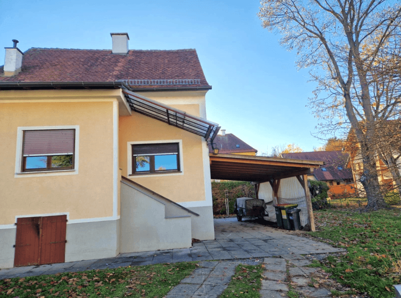 Seitenansicht des Hauses mit Carport und Fenstern mit Rollläden unter blauem Himmel.