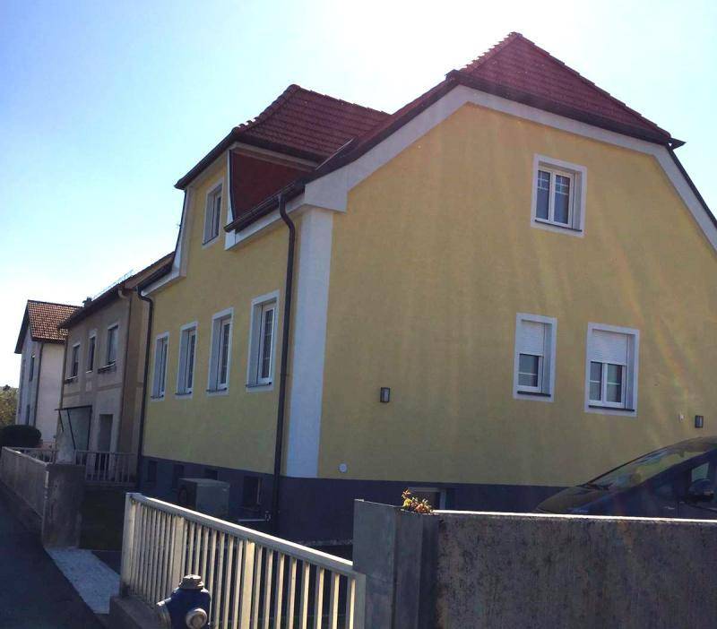 Seitenansicht eines gelben Hauses mit rotem Satteldach, weißen Fenstern und Rollläden unter blauem Himmel.