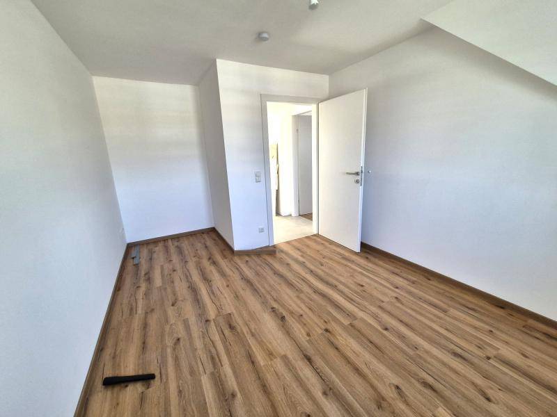Leeres Zimmer mit Laminatboden und weißen Wänden, mit offener Tür zu einem angrenzenden Bereich.