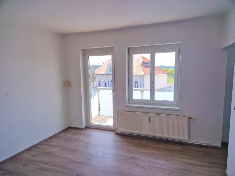 Helles Zimmer mit Laminatboden, großem Fenster und Zugang zu einem kleinen Balkon.