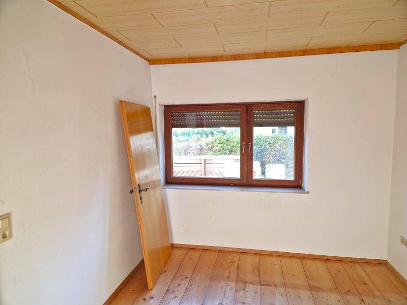 Leerer Raum mit Holzboden, Fenster mit Rollladen und Blick ins Grüne.