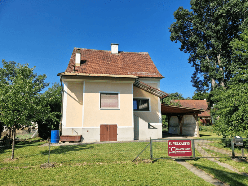 Vorderansicht des Hauses mit Garten, Zaun und einem "Zu Verkaufen" Schild.