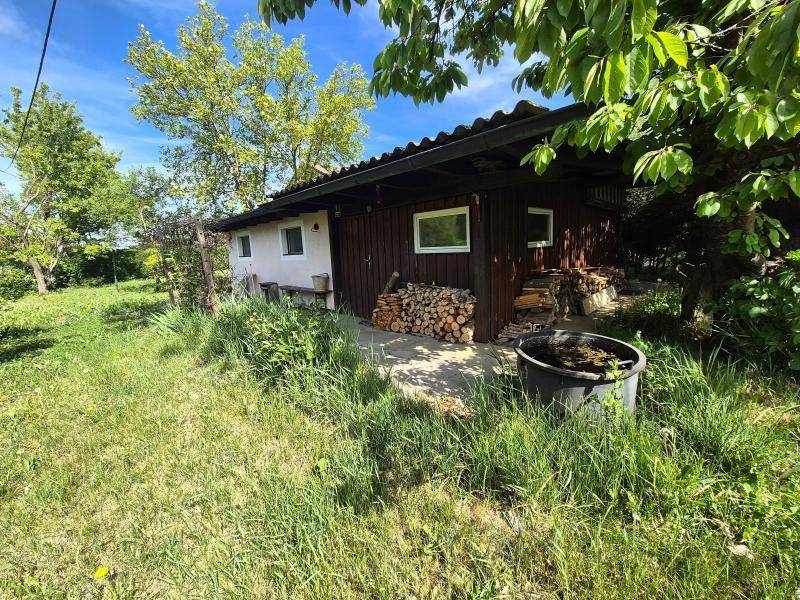 Rustikales Holzgebäude im Garten mit gestapeltem Brennholz und umgebender Natur.