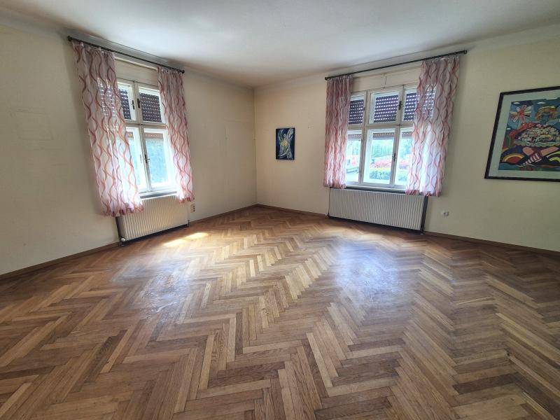 Geräumiges Zimmer mit Fischgrätparkett, zwei Fenstern und Heizkörpern unter den Fenstern.