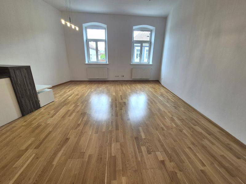 Heller Wohnbereich mit Holzboden, weißen Wänden und zwei großen Bogenfenstern, die viel Licht hereinlassen.