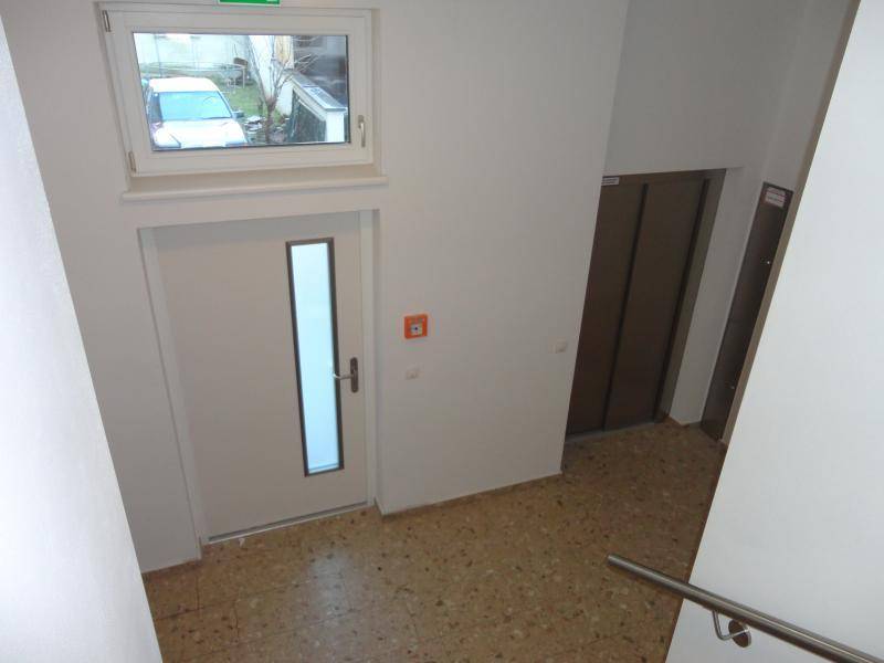 Treppenhausbereich mit Fenster, Zugangstür und einem sichtbaren Aufzug im Hintergrund.