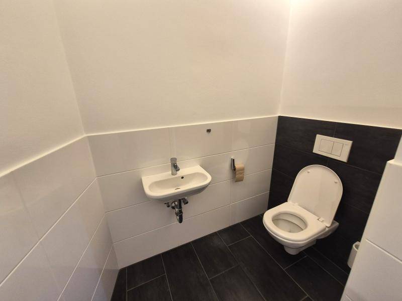Gäste-WC mit dunklen Bodenfliesen, weißen Wandfliesen, wandhängendem WC und kleinem Handwaschbecken.