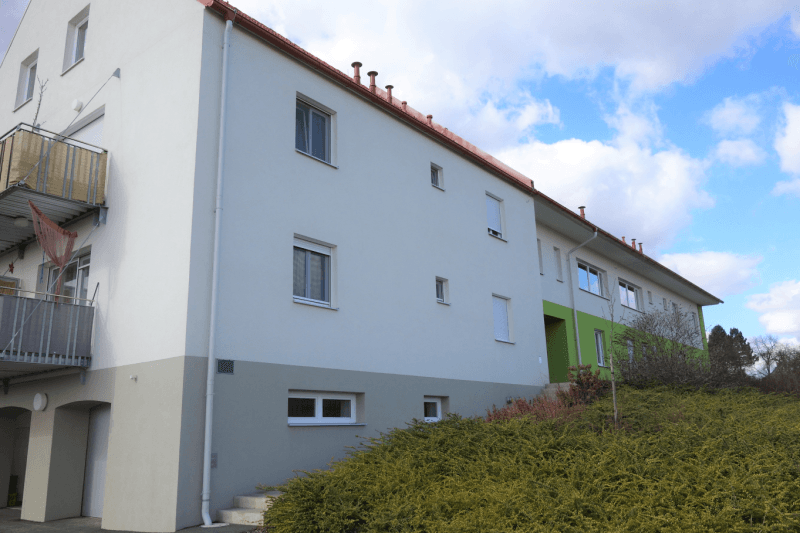 Ansicht des Mehrfamilienhauses mit weißer und grauer Fassade sowie Balkonen.